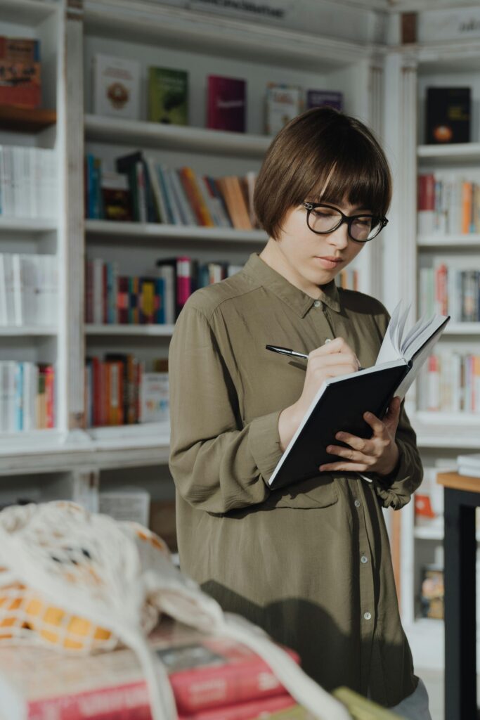 Young woman with glasses writing in a notebook at a bookstore surrounded by shelves of books.