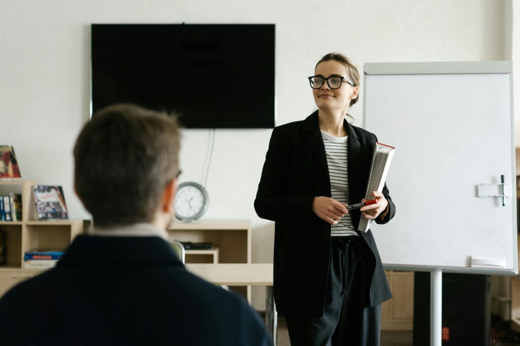 pexels-photo-6981013-6981013 Caucasian female teacher in black blazer holding books, presenting to a student in a classroom.