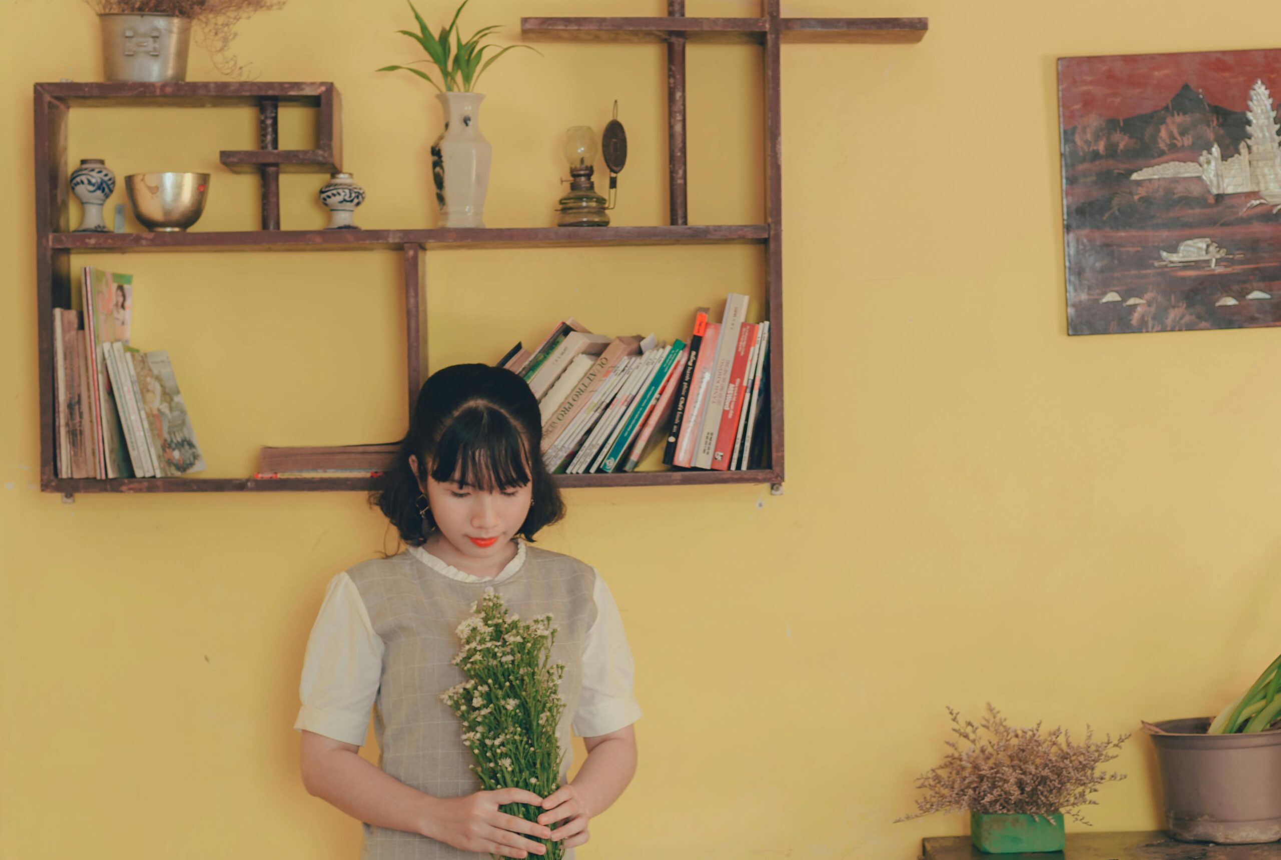 A young woman holding flowers in a stylish room with books and decor.