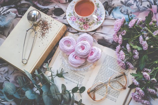 Delicate flatlay with pink pastries, tea, flowers, book, and glasses for a romantic setting.