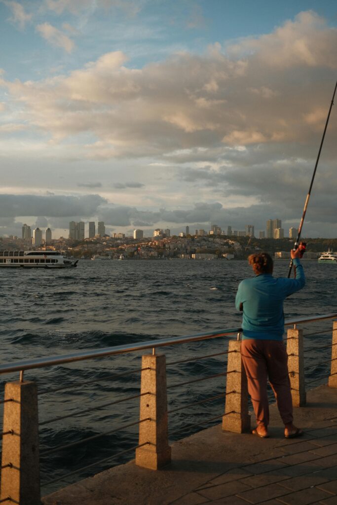 pexels-photo-18432645-18432645 A man fishing on a pier with a city skyline in the background during sunset, creating a serene urban scene.