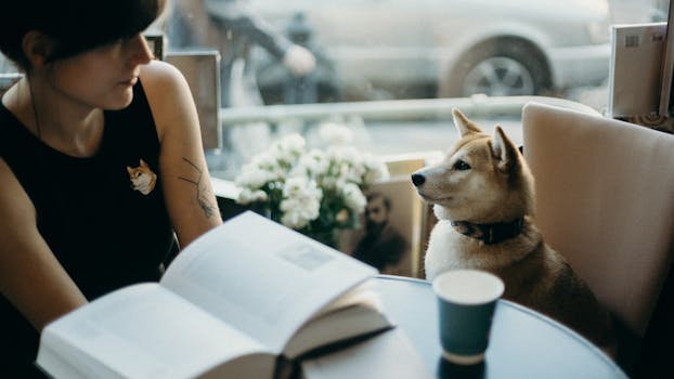 A woman enjoys a book at a cafe with her Shiba Inu dog by her side.