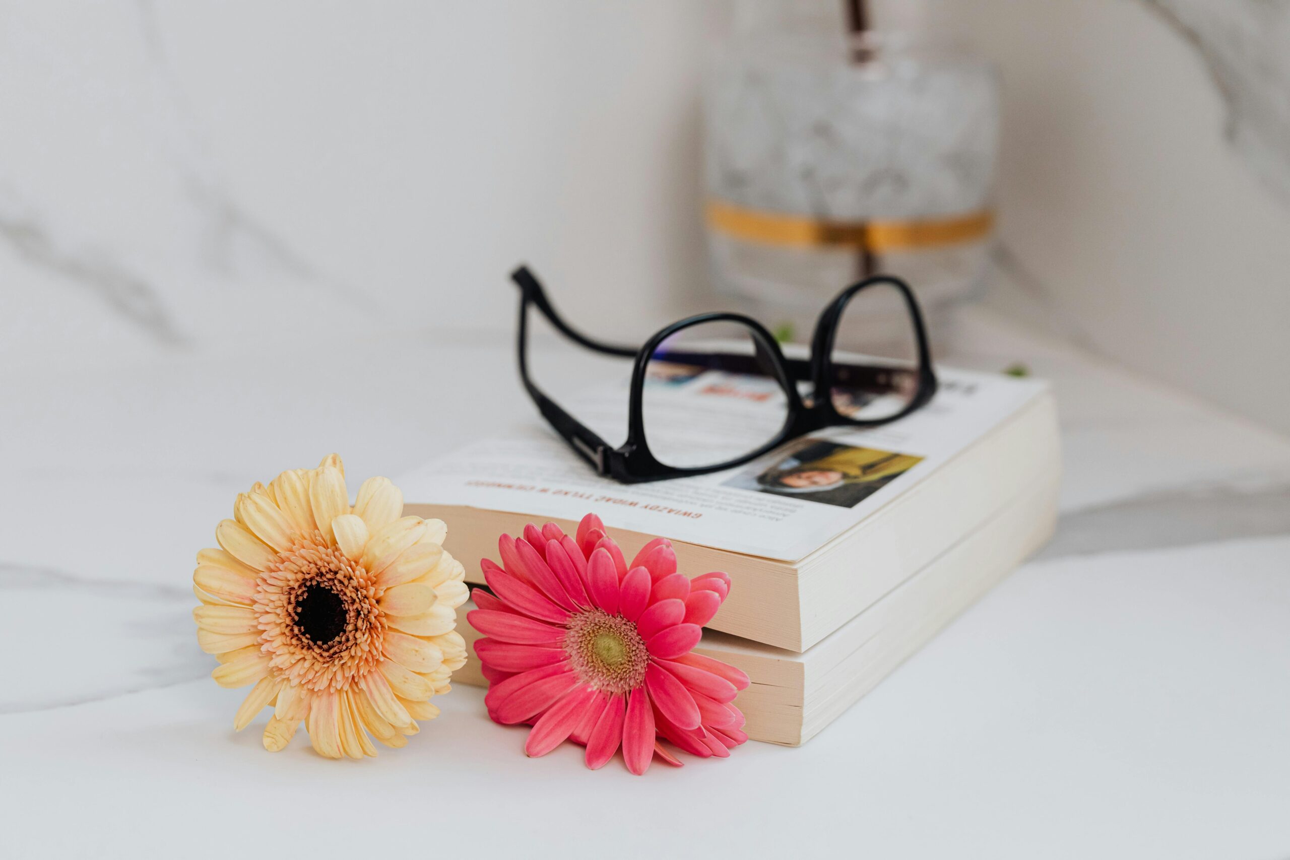 An elegant still life of gerbera flowers, eyeglasses, and books on a marble surface.