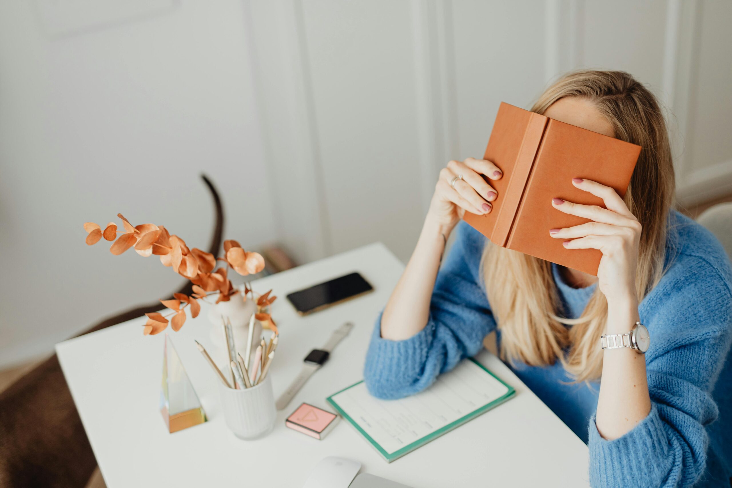 A woman in a blue sweater reading a book at a modern workspace indoors.