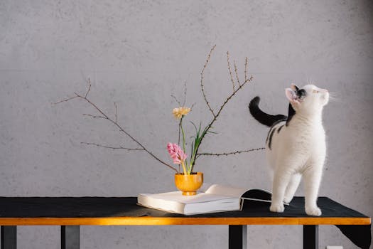 A curious black and white cat explores near a vase with flowers on a book, creating a whimsical indoor scene.
