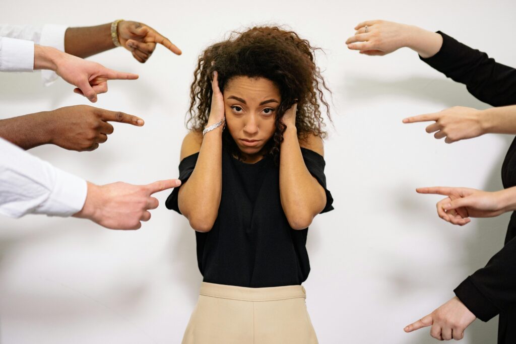 pexels-photo-7640496-7640496 Stressed woman with hands on ears surrounded by pointing fingers, illustrating pressure.