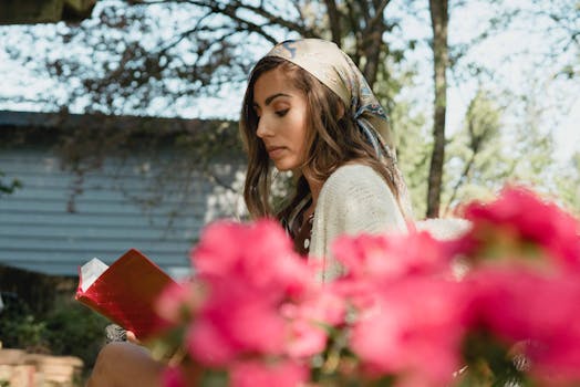 Young woman reading a book outdoors, surrounded by vibrant flowers, wearing a colorful scarf.