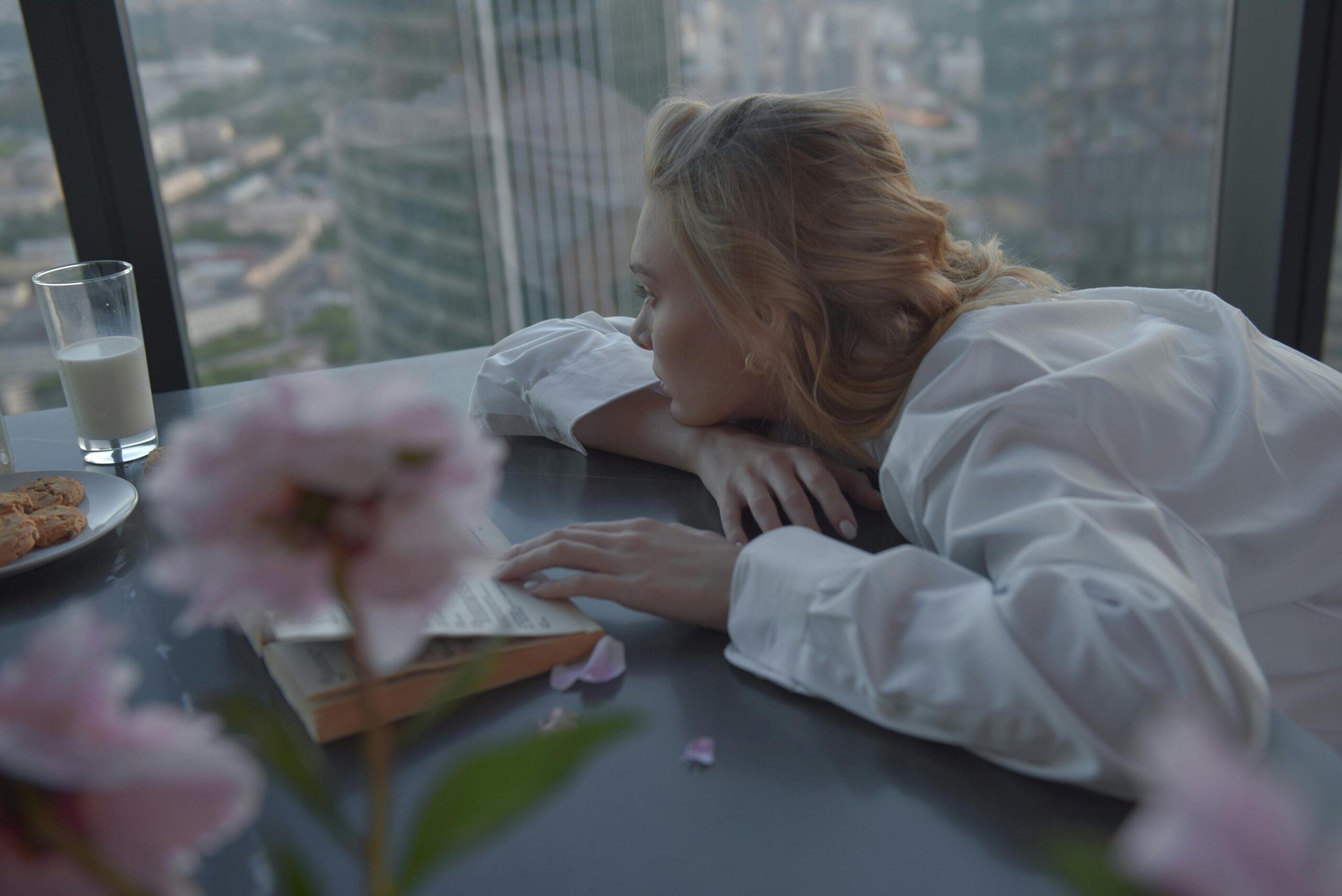 Woman in serene moment, reading by a large window with urban cityscape view, enjoying milk and cookies.
